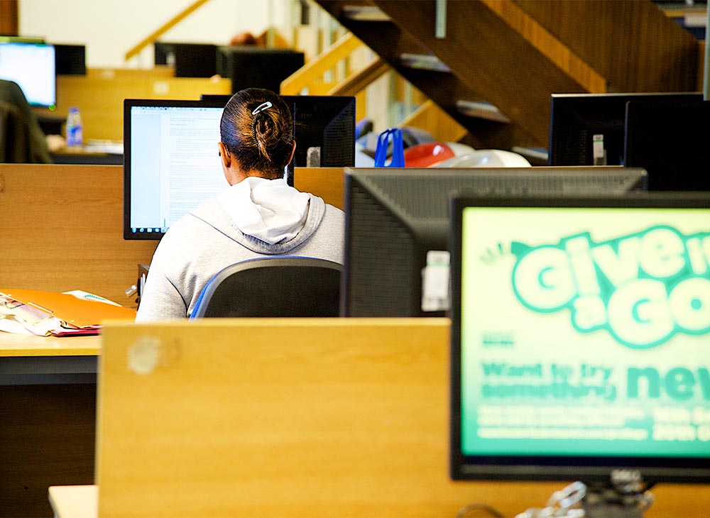 Image of a computer cluster room, with several active screens. You can see one student engaged with their work on screen.